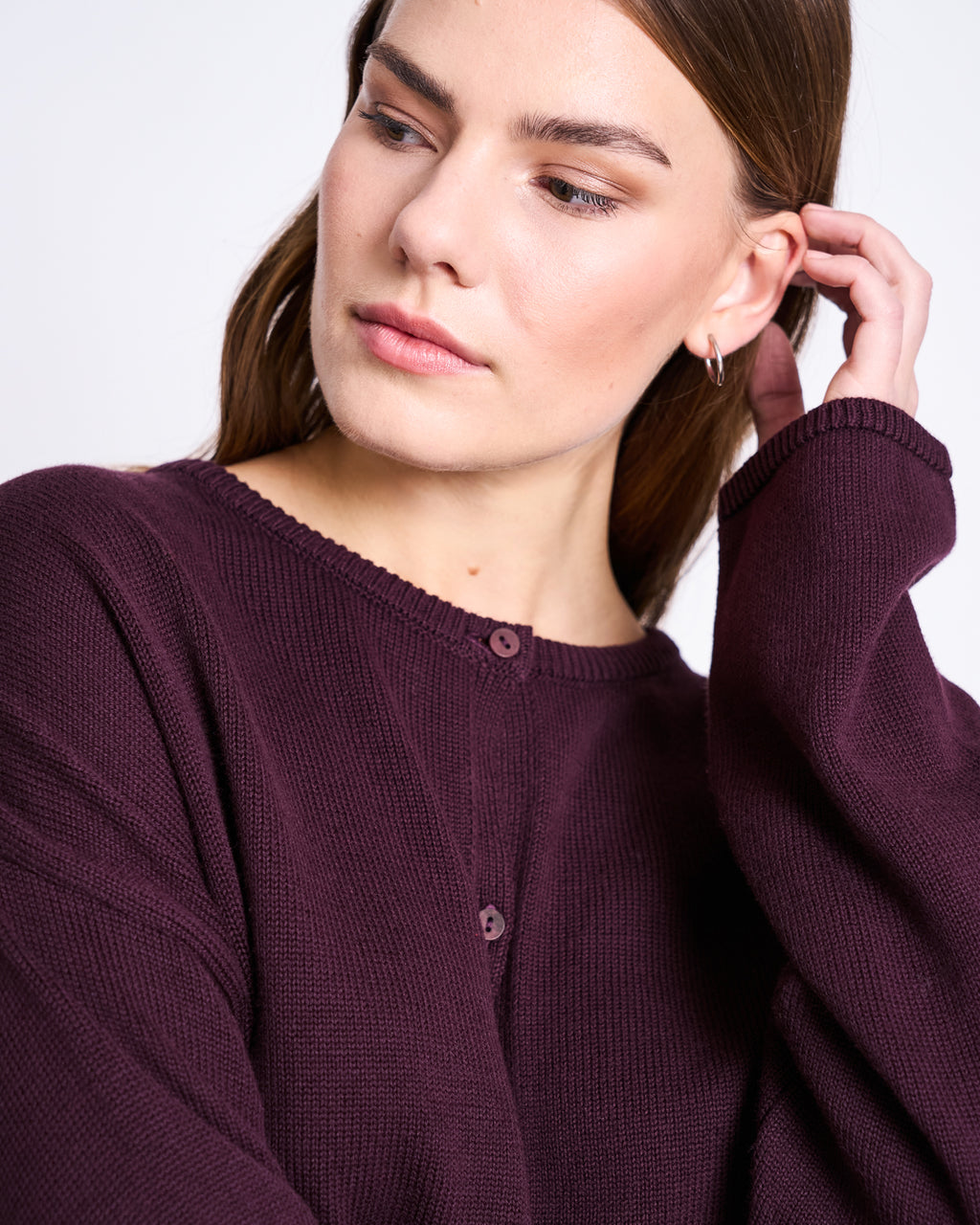 A woman with straight brown hair wears the jan-n-june LIGHT CARDIGAN ASAS CHERRY LACQUER GOTS and small hoop earrings, looking to the side while touching her hair against a plain, light background.