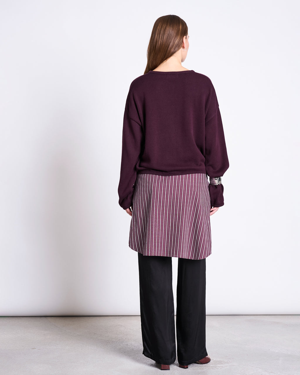 A woman faces away from the camera, wearing the jan-n-june LIGHT CARDIGAN ASAS CHERRY LACQUER GOTS, a purple striped skirt over black wide-leg pants, and brown shoes against a plain white background.