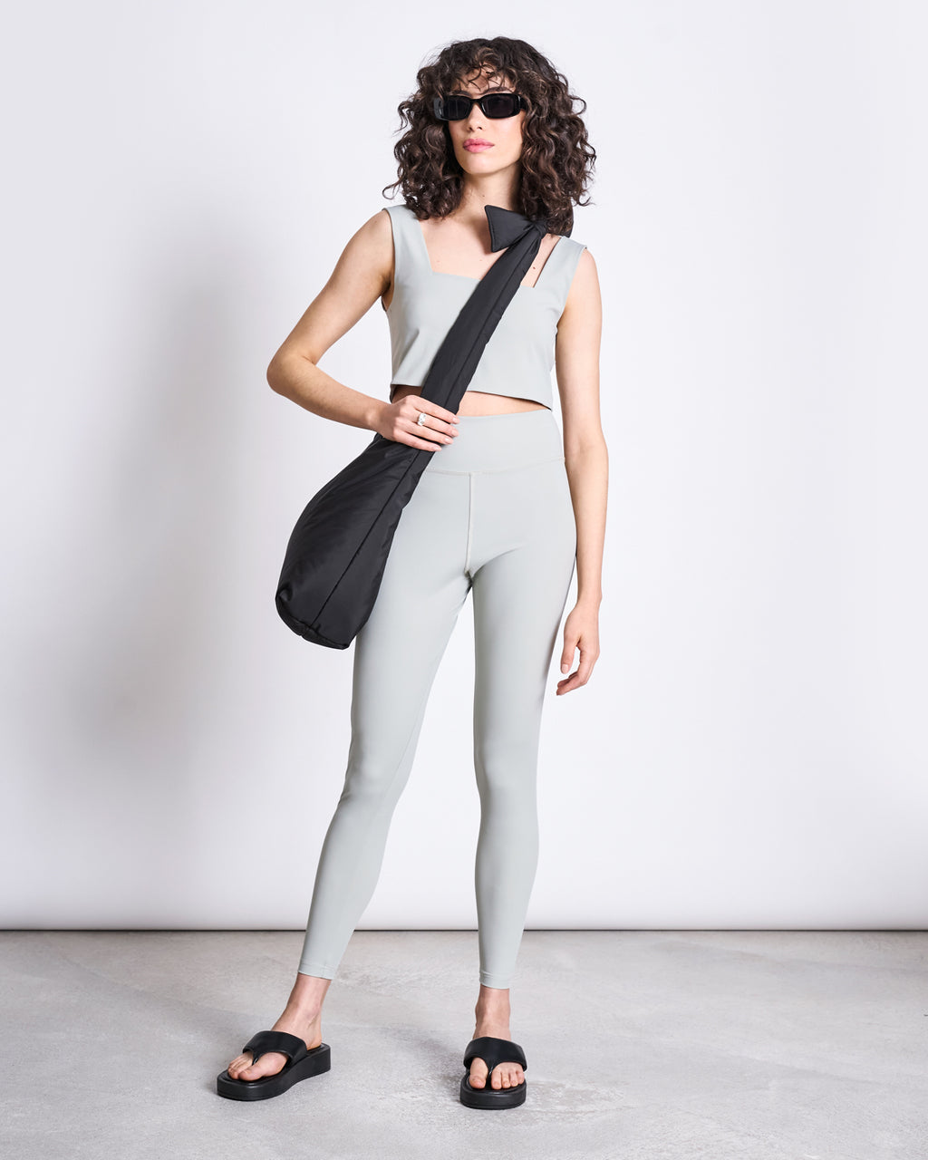 A woman with curly hair wearing sunglasses, a light gray sleeveless crop top, jan-n-june ACTIVE LEGGINGS HALLIN GLACIAL GREEN, black platform sandals, and a black shoulder bag stands against a plain white background.