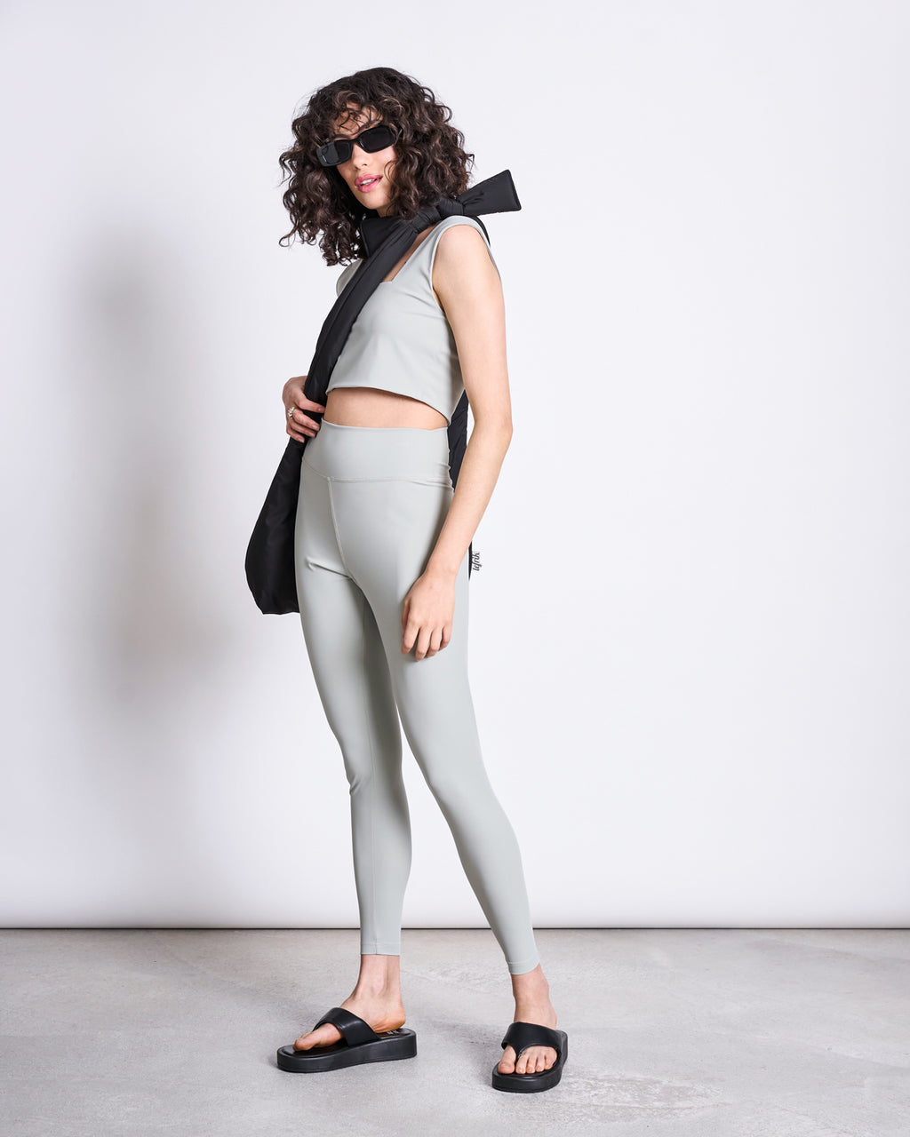 A woman with curly hair wears black sunglasses, a light gray crop top, jan-n-june ACTIVE LEGGINGS HALLIN in Glacial Green made from recycled polyamide, black sandals, and a black scarf over her shoulder as she stands sideways against a plain background.