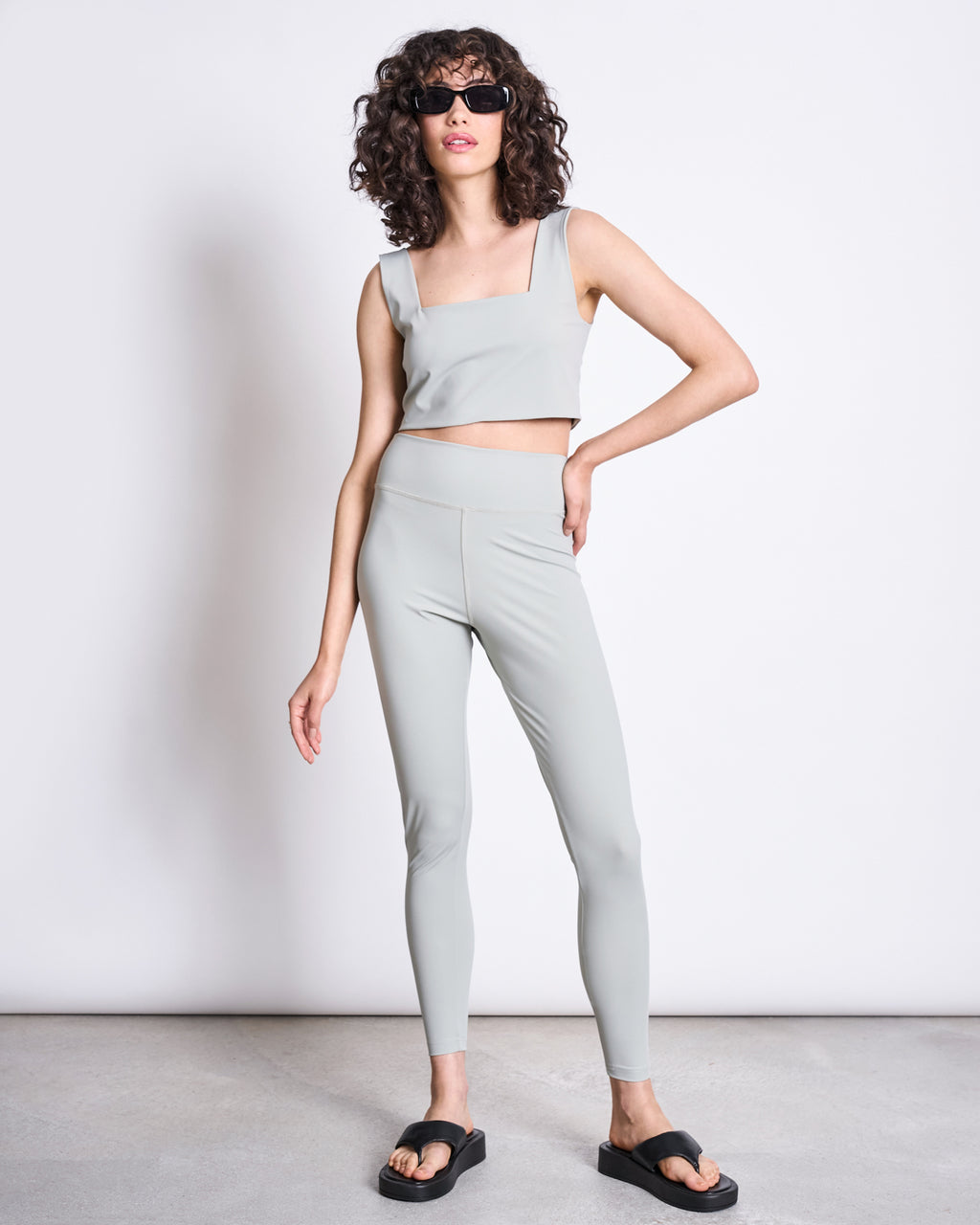A woman with curly hair wears sunglasses, a light gray crop top, and jan-n-june ACTIVE LEGGINGS HALLIN GLACIAL GREEN made from recycled polyamide, paired with black slide sandals, standing confidently against a plain white background.