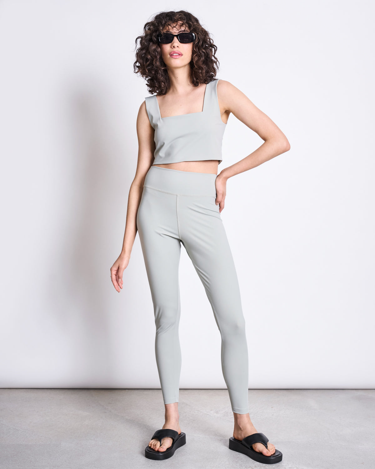 A woman with curly hair wears sunglasses, a light gray crop top, and jan-n-june ACTIVE LEGGINGS HALLIN GLACIAL GREEN made from recycled polyamide, paired with black slide sandals, standing confidently against a plain white background.
