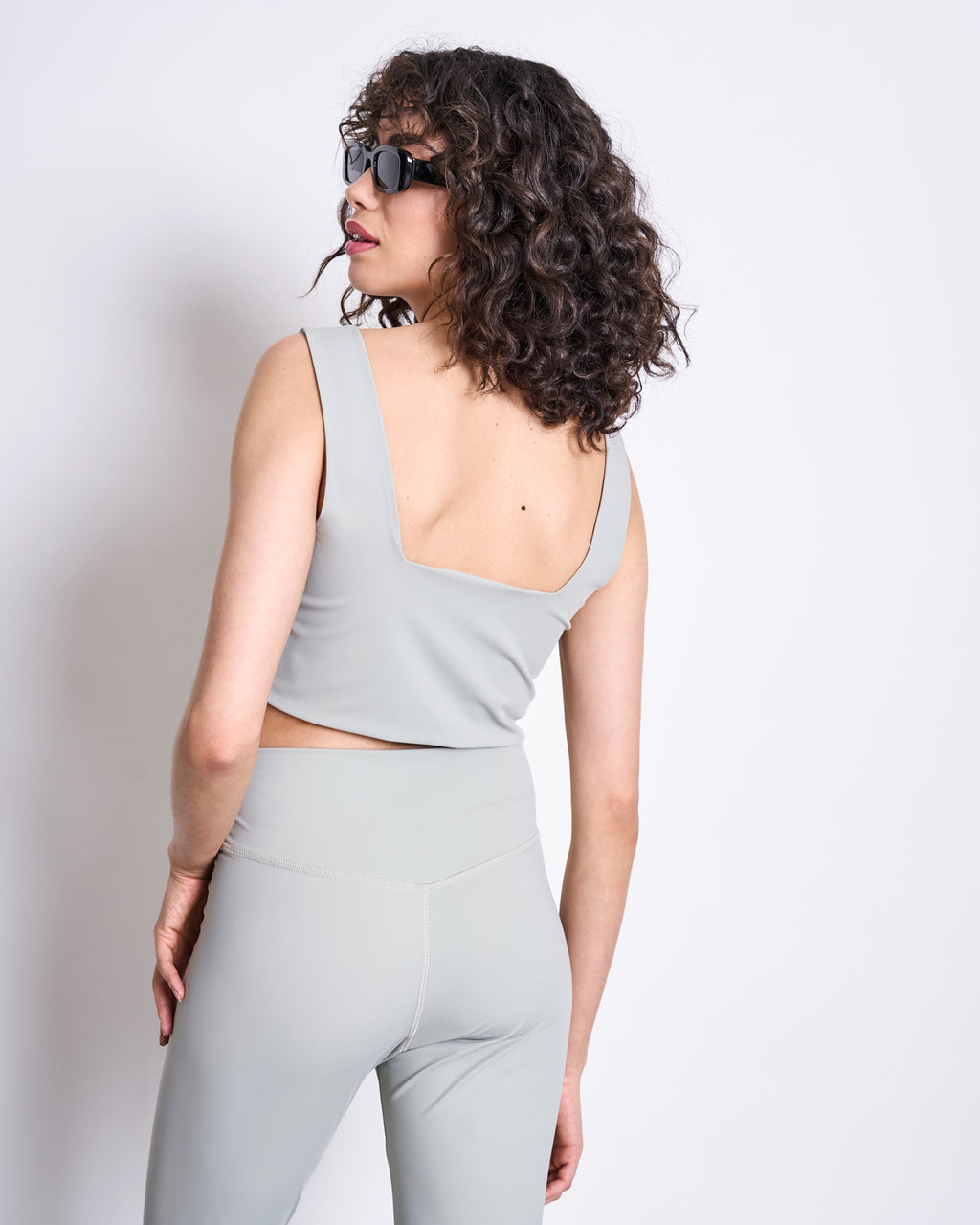 A person with curly hair and sunglasses poses with their back slightly turned, wearing jan-n-junes ACTIVE LEGGINGS HALLIN GLACIAL GREEN and a matching light gray sleeveless crop top made from recycled polyamide against a plain background.