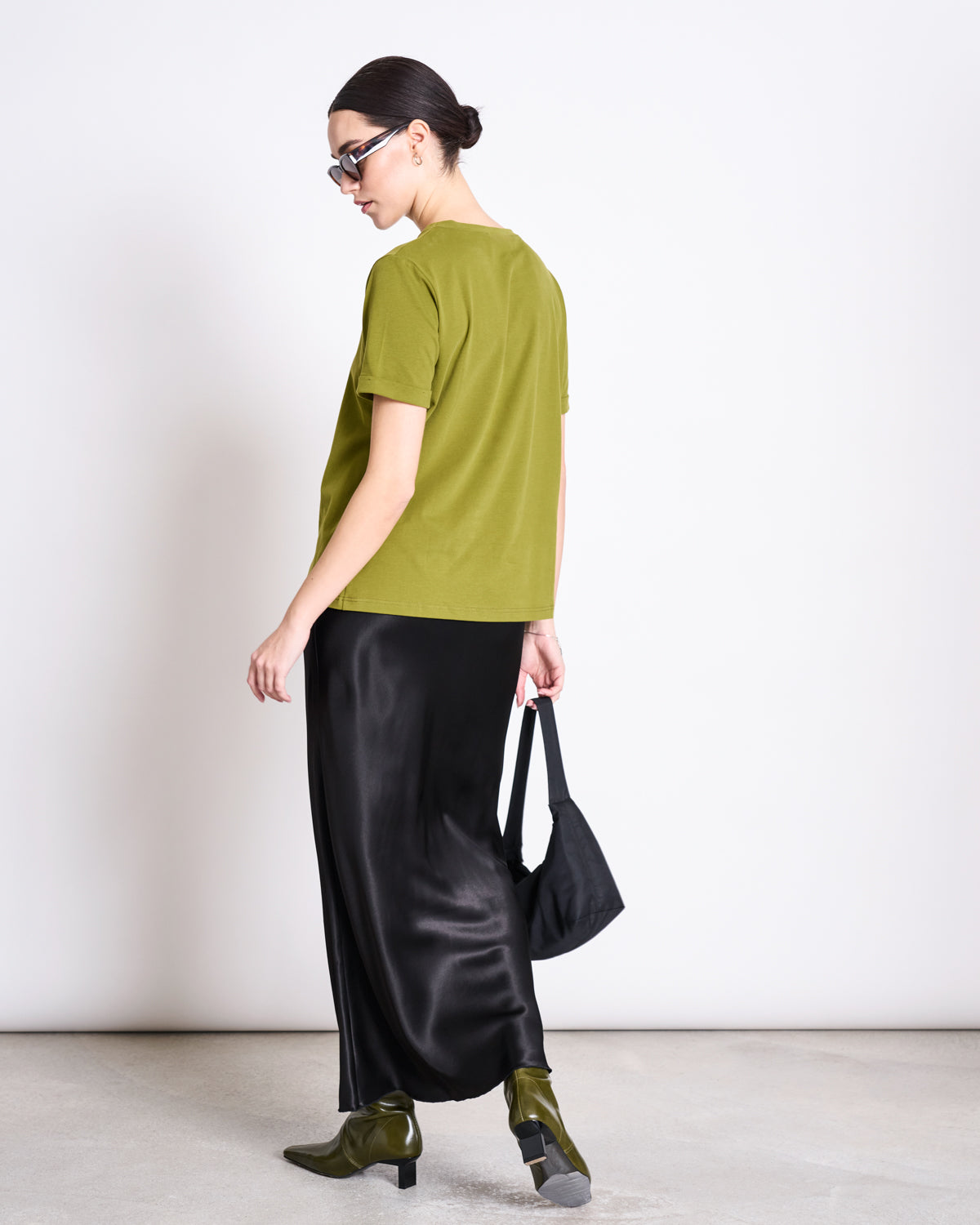 A woman in sunglasses and the jan-n-june BRUSHED T-SHIRT BOY WOODY OLIVE GOTS, paired with a long black satin skirt, olive ankle boots, and a black handbag stands facing away against a plain white background.