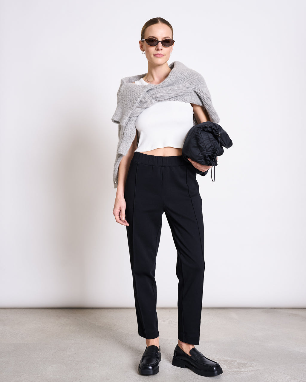 A woman stands indoors against a plain white background wearing sunglasses, a gray sweater over her shoulders, a white cropped top, and jan-n-junes high-waisted PANTS KAMA BLACK GOTS, holding a black bag.