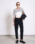 A woman stands indoors against a plain white background wearing sunglasses, a gray sweater over her shoulders, a white cropped top, and jan-n-junes high-waisted PANTS KAMA BLACK GOTS, holding a black bag.
