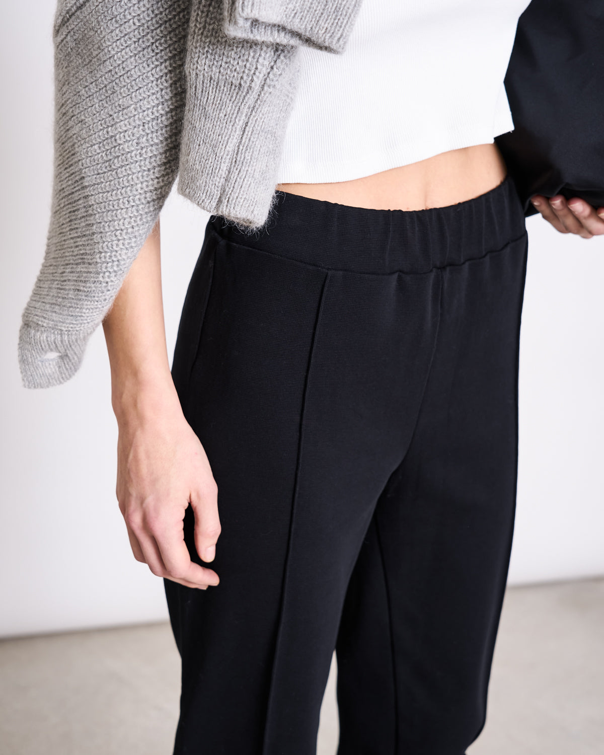 A person wearing jan-n-june’s PANTS KAMA BLACK GOTS, paired with a white top and a gray sweater draped over their shoulder, stands indoors with their hand at their side.