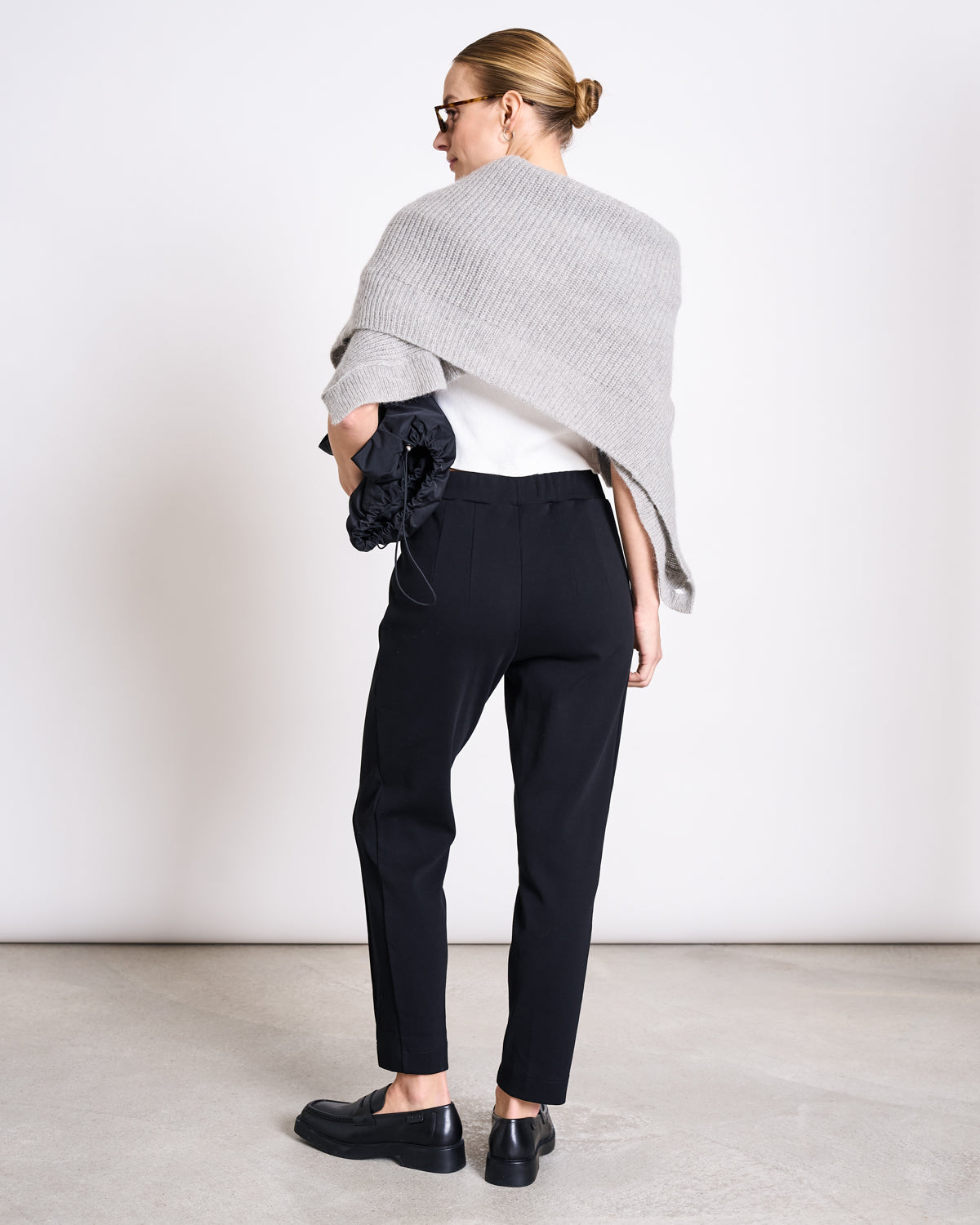 A woman, with her hair in a neat bun and sunglasses, wears jan-n-junes high-waisted PANTS KAMA BLACK GOTS, black loafers, a white top, a gray wrap over her shoulders, and holds a black clutch against a plain light background.
