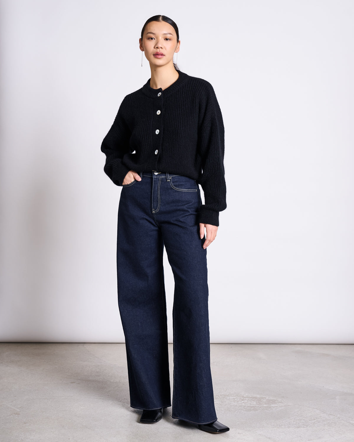 A woman stands confidently against a plain background, wearing jan-n-june’s WIDE LEG LOWRISE JEANS TACNA DARK BLUE DENIM GOTS and a black buttoned cardigan. Her hair is tied back, one hand in her pocket, and she wears black shoes.