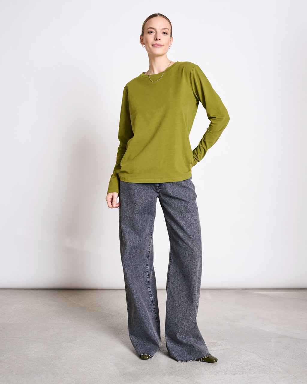 A woman stands in a minimalist room wearing the jan-n-june BRUSHED LONGSLEEVE BOY WOODY OLIVE GOTS top, paired with loose-fit wide-leg gray jeans. She smiles slightly, one hand on her hip, and wears green pointed shoes and a simple necklace.