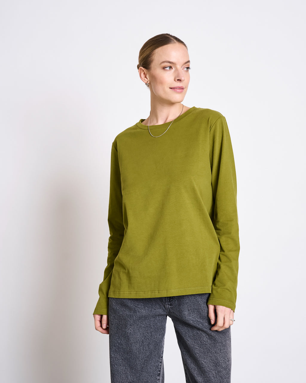 A woman wears the jan-n-june BRUSHED LONGSLEEVE BOY WOODY OLIVE GOTS shirt with dark gray jeans, standing against a white background and smiling gently as she looks slightly to her left.