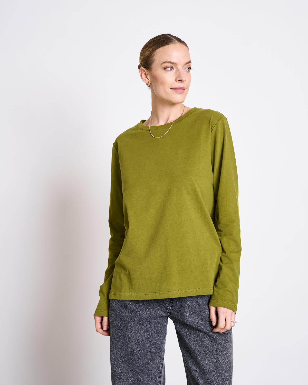 A woman wears the jan-n-june BRUSHED LONGSLEEVE BOY WOODY OLIVE GOTS shirt with dark gray jeans, standing against a white background and smiling gently as she looks slightly to her left.