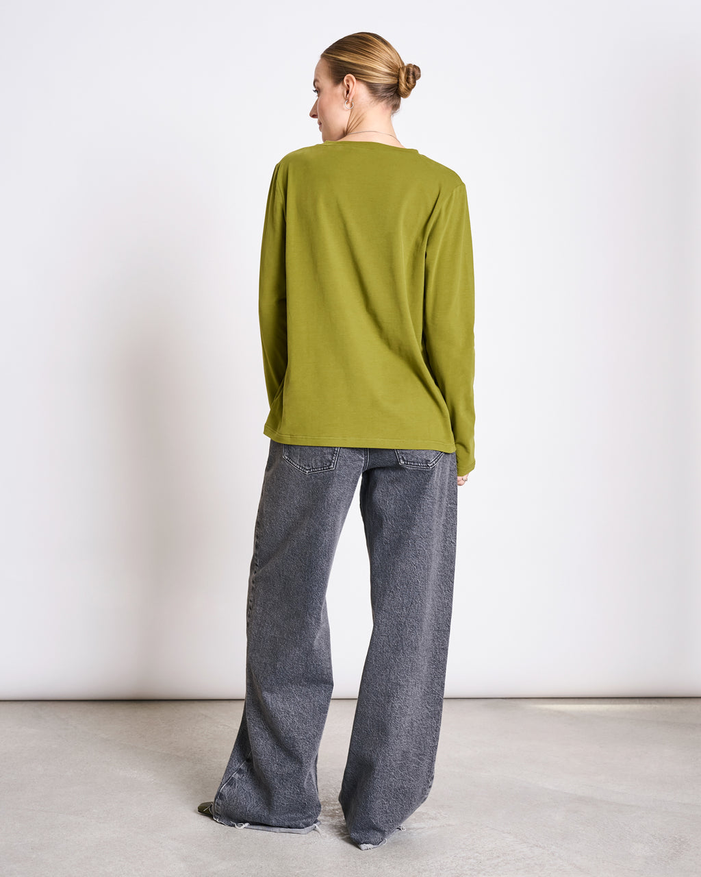 A person with tied back hair stands indoors facing away, wearing the jan-n-june BRUSHED LONGSLEEVE BOY WOODY OLIVE GOTS and boyfriend style gray jeans, against a plain light background.