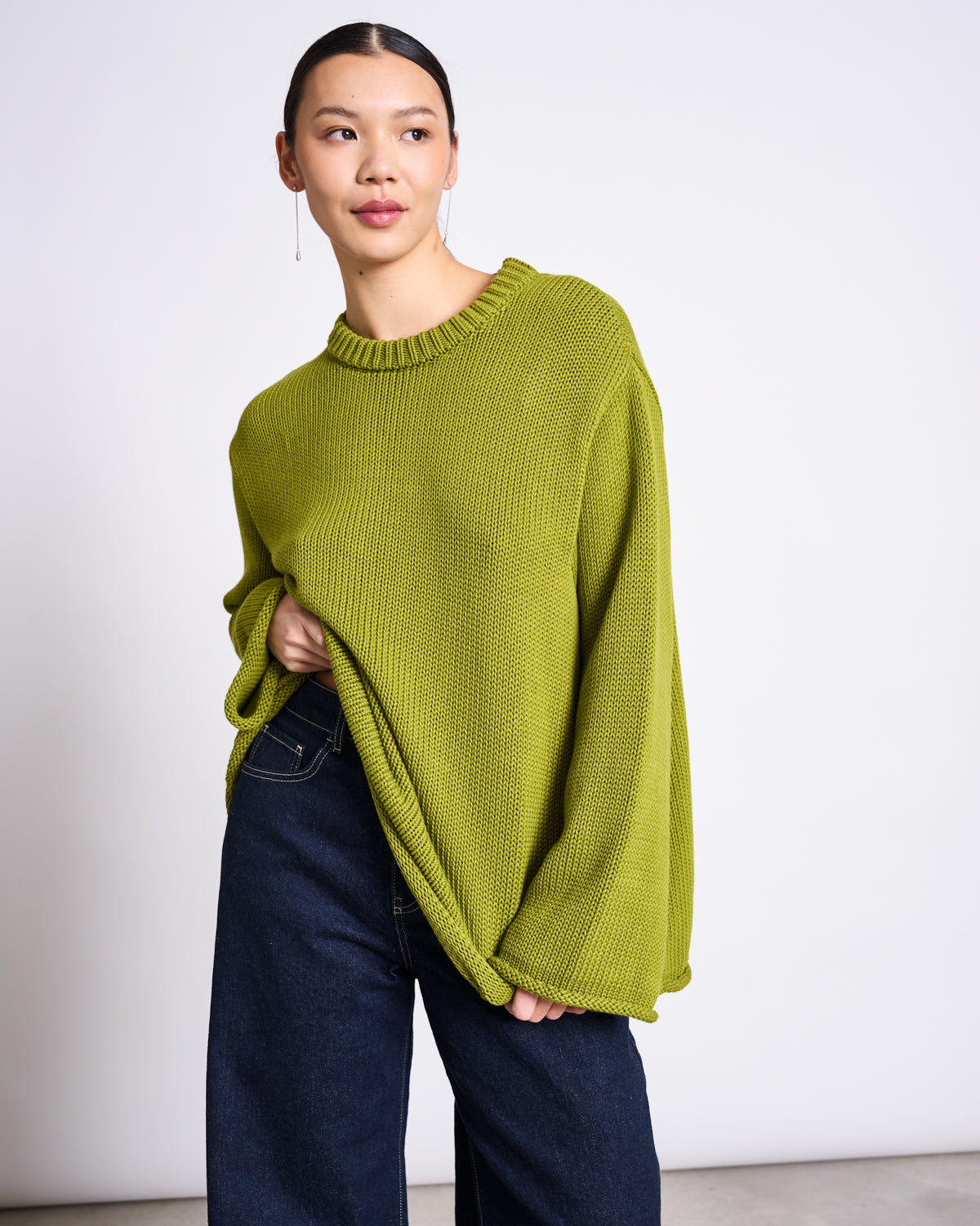 A person wears the jan-n-june JUMPER SONA MOSSY PEAR GOTS, an oversized bright green organic cotton sweater, paired with dark wide-leg jeans. They stand against a white background, looking aside while holding the sweaters hem.