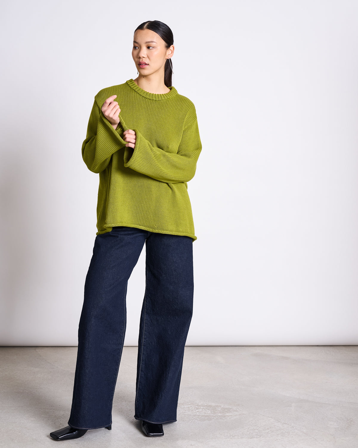 A person stands indoors wearing the jan-n-june JUMPER SONA MOSSY PEAR GOTS—a bright green, oversized knit sweater made from GOTS certified organic cotton—paired with wide-leg dark jeans and black shoes against a plain light background.