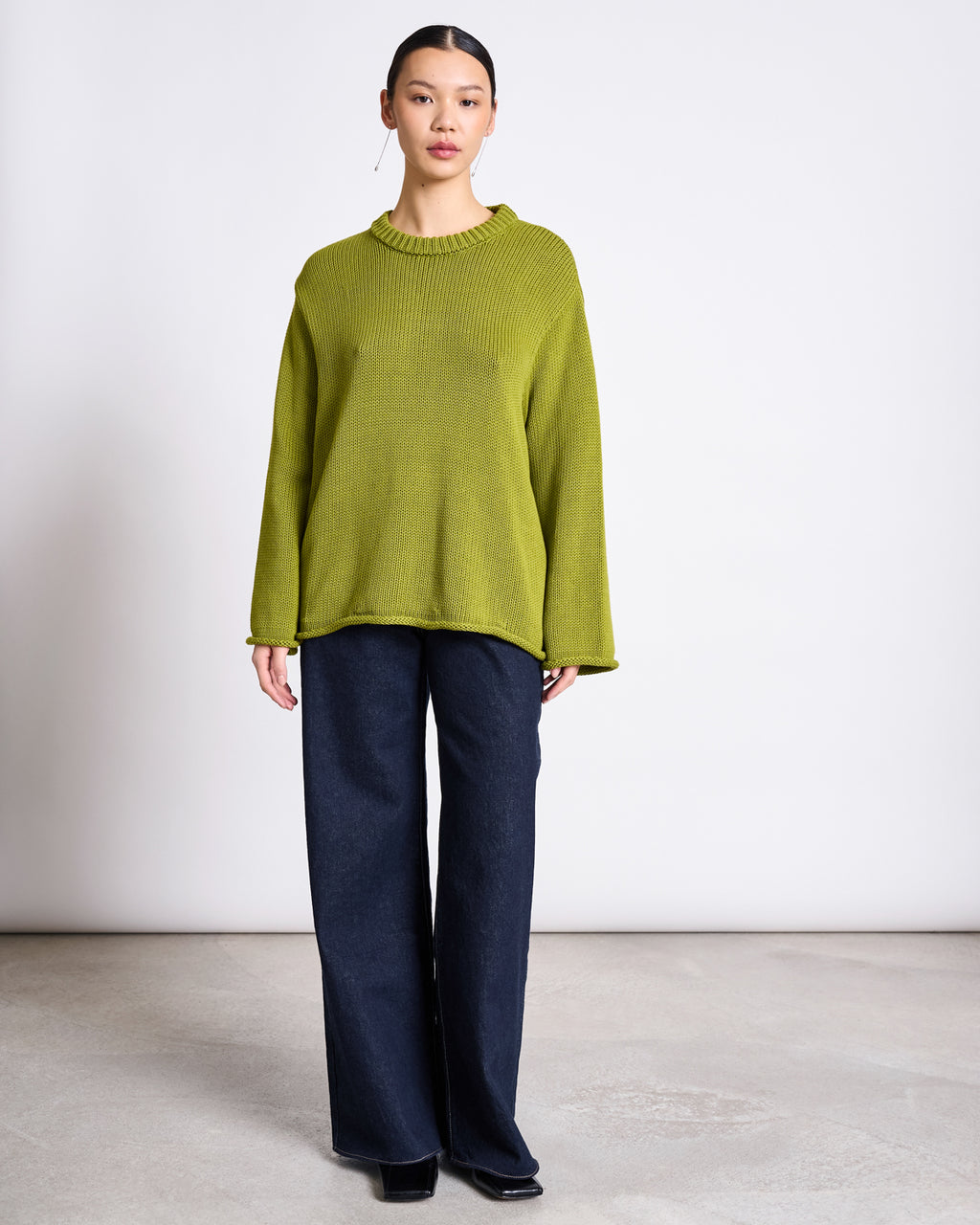 A person stands indoors against a plain white wall, wearing the jan-n-june JUMPER SONA MOSSY PEAR GOTS, an oversized light green organic cotton sweater, paired with wide-leg dark blue jeans and black shoes.