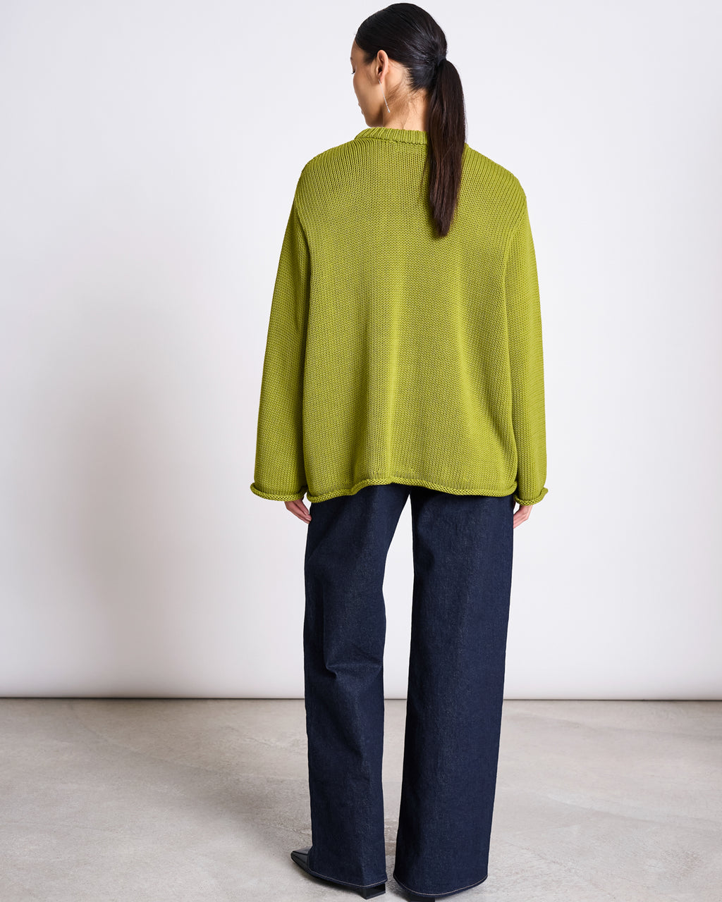 A person with long dark hair in a ponytail faces away, wearing the jan-n-june JUMPER SONA MOSSY PEAR GOTS, an oversized bright green organic cotton sweater, with wide-leg dark jeans on a light floor against a white wall.