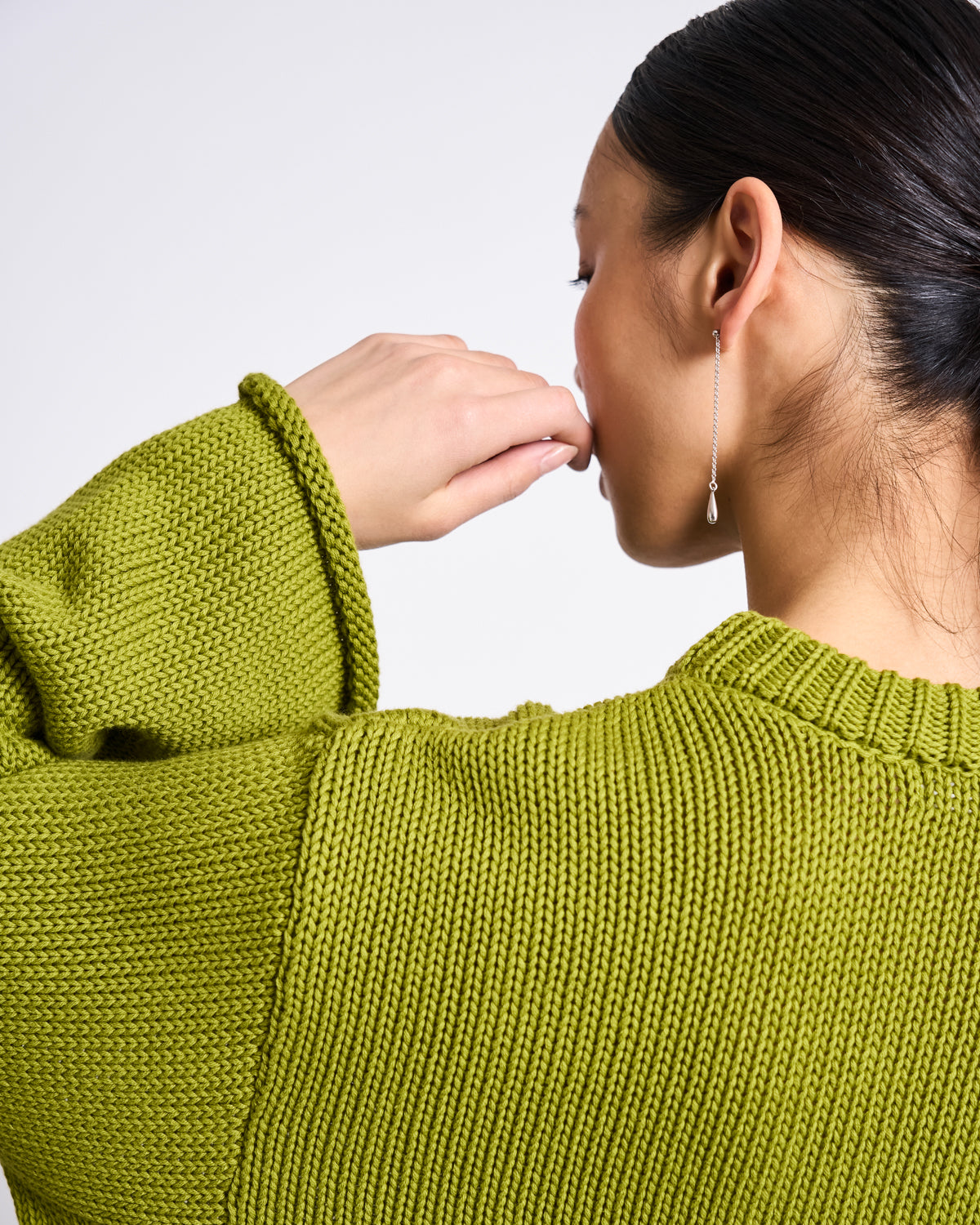 A woman with dark hair in a low bun wears the jan-n-june JUMPER SONA MOSSY PEAR GOTS, an oversized green organic cotton sweater, and a long earring. She is seen from behind and in profile, her hand near her face against a plain background.