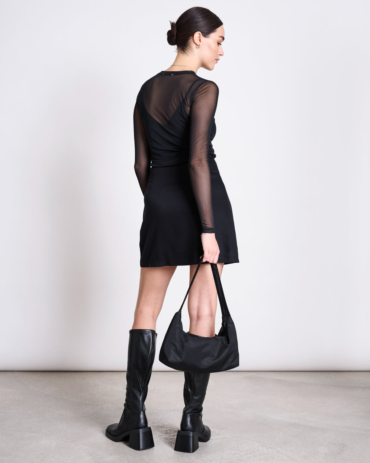 A woman with her hair in a low bun wears the jan-n-june MESH LONGSLEEVE DORA BLACK, paired with a black skirt, knee-high black boots, and holding a black handbag against a plain light background.