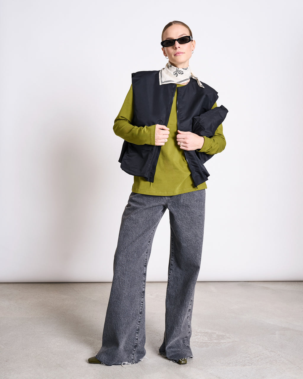 Against a light background, a person wears black sunglasses, a black vest over the jan-n-june BRUSHED LONGSLEEVE BOY WOODY OLIVE GOTS top, a white collared shirt, wide-leg gray jeans, and pointed shoes in a modern, fashion-forward style.