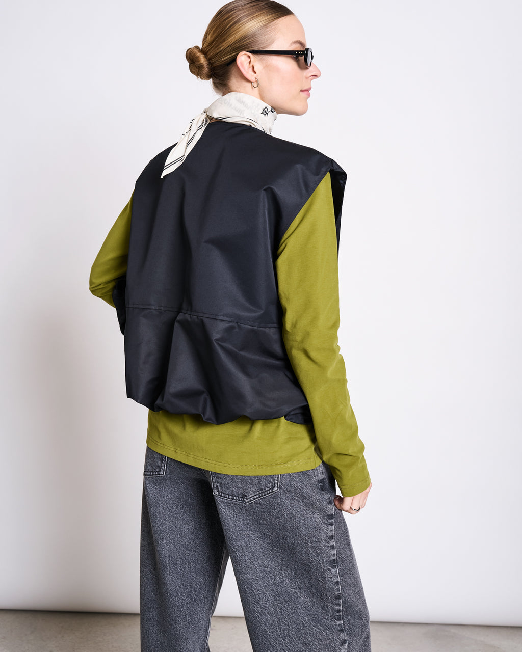 A person with light brown hair in a bun, black glasses, a white patterned scarf, black vest, and loose gray jeans wears the jan-n-june BRUSHED LONGSLEEVE BOY WOODY OLIVE GOTS shirt, facing away from the camera against a white background.