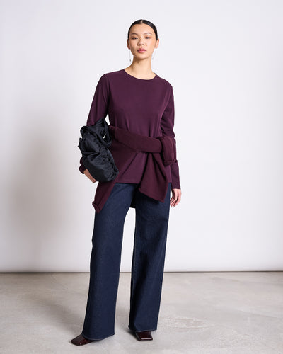 A woman wears the jan-n-june BRUSHED LONGSLEEVE BOY CHERRY LACQUER GOTS top in burgundy, paired with a matching sweater at her waist, loose dark blue wide-leg jeans, pointed burgundy shoes, and holds a black puffer jacket.