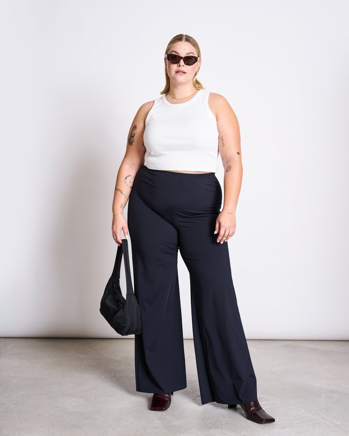 A person with blonde hair and sunglasses stands against a plain background, wearing a jan-n-june ORGANIC COTTON TANK TOP PORTO WHITE GOTS, black wide-leg pants, dark shoes, and holding a black bag in their left hand.