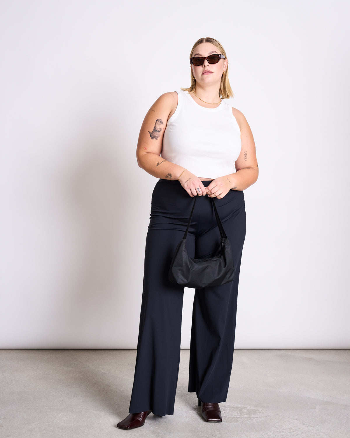 A woman stands against a plain background wearing jan-n-june’s WIDE PANTS MARLENE BLACK, styled with sunglasses, a white sleeveless top, burgundy shoes, and holding a black handbag in both hands.