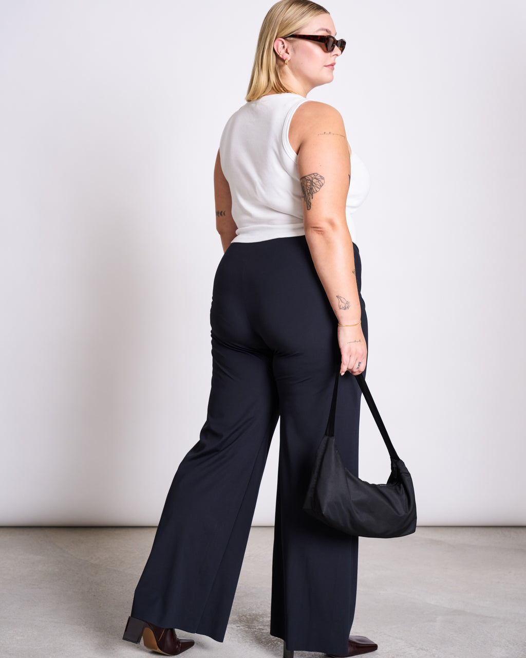 A woman with blonde hair, sunglasses, and visible arm tattoos wears a white sleeveless top and jan-n-junes WIDE PANTS MARLENE BLACK made from sustainable fabric, facing away while holding a black handbag at her side.