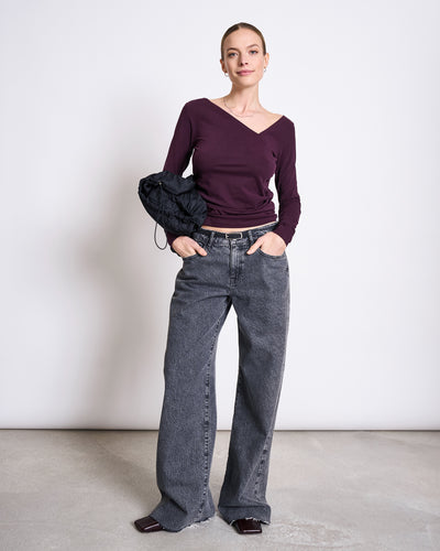 A woman with her hair pulled back wears jan-n-june’s ASYMMETRICAL LONGSLEEVE CASMA CHERRY LACQUER GOTS top, loose dark gray jeans, square-toed shoes, and holds a black bag against a plain background.