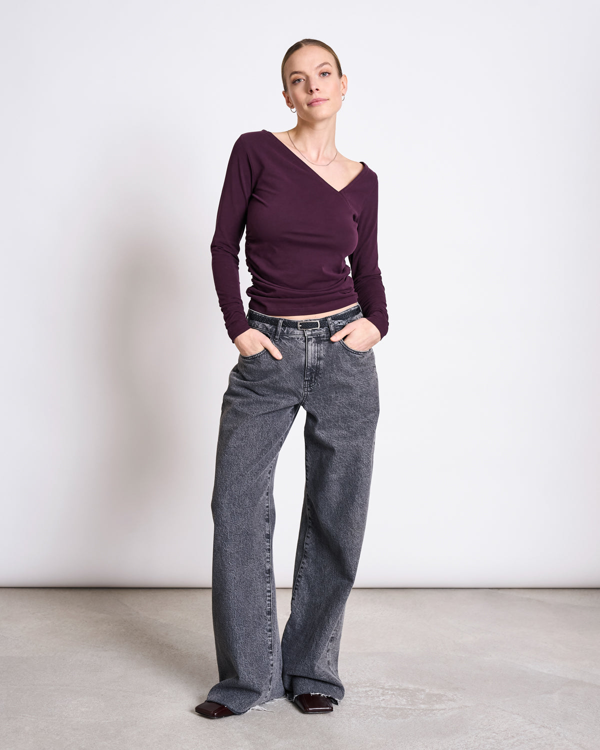 A woman stands against a plain white wall, wearing the jan-n-june ASYMMETRICAL LONGSLEEVE CASMA CHERRY LACQUER GOTS top and loose gray jeans with a belt. Her hands are in her pockets as she looks confidently at the camera.