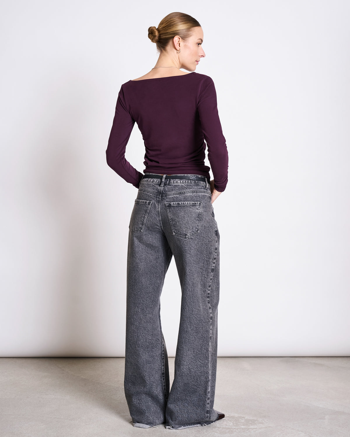 A woman with blonde hair in a bun wears the jan-n-june ASYMMETRICAL LONGSLEEVE CASMA CHERRY LACQUER GOTS and loose gray jeans, standing with her back to the camera against a plain white wall.