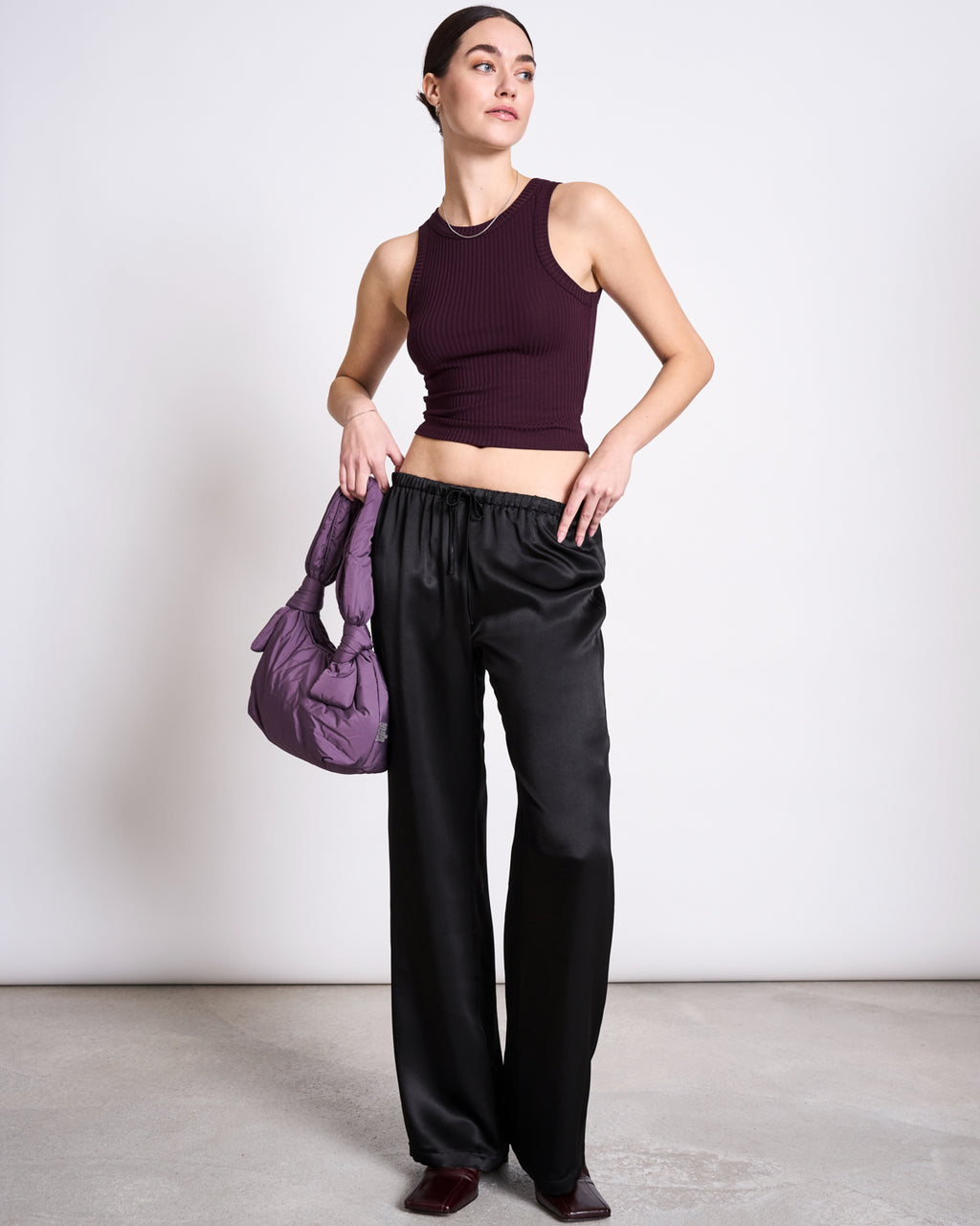 A woman stands indoors wearing the jan-n-june LYOCELL RIB TANK TOP PORTO CHERRY LACQUER, loose black satin pants, and dark shoes. She holds a padded purple handbag and gazes to the side against a plain light background.