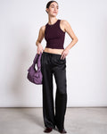 A woman stands indoors wearing the jan-n-june LYOCELL RIB TANK TOP PORTO CHERRY LACQUER, loose black satin pants, and dark shoes. She holds a padded purple handbag and gazes to the side against a plain light background.