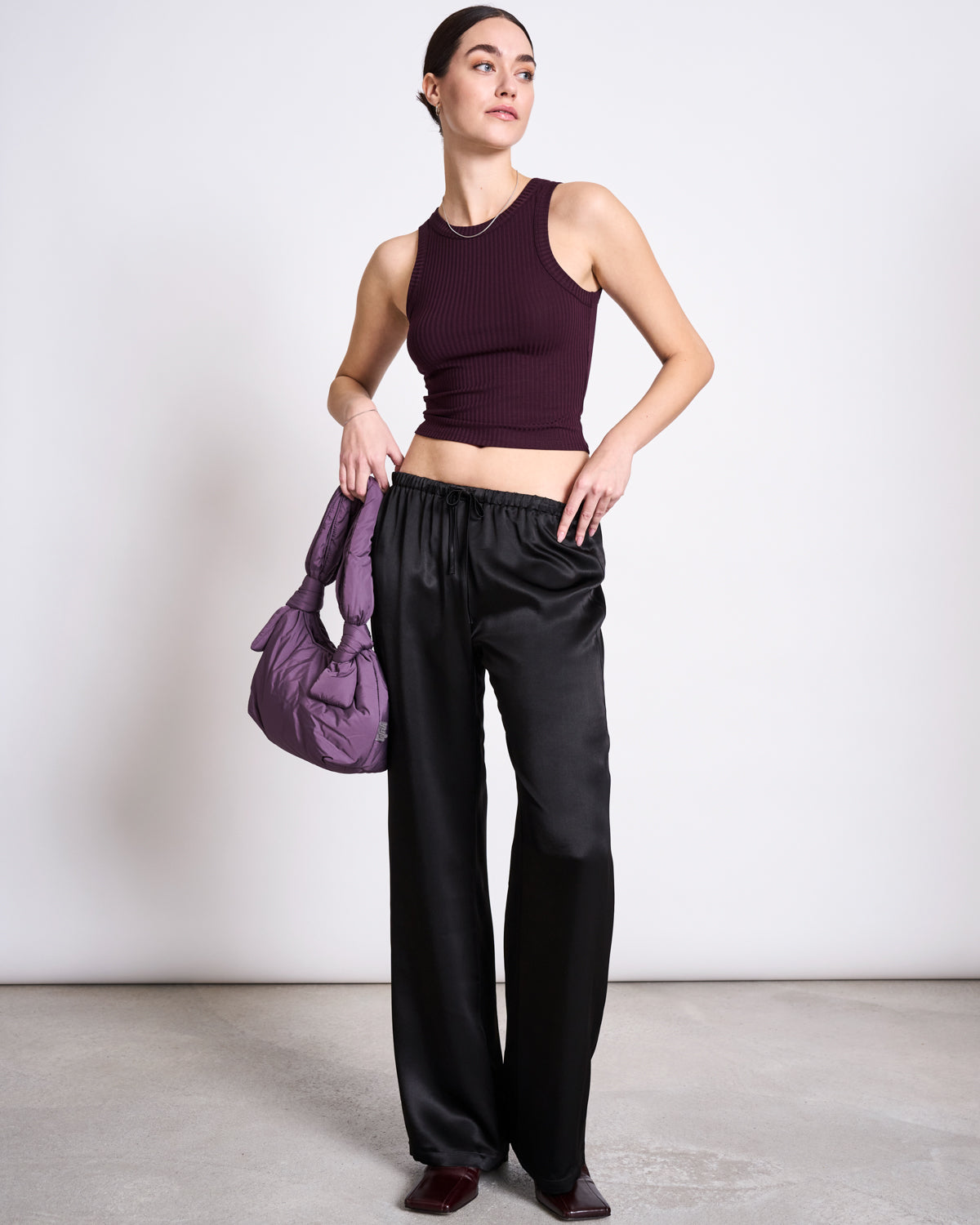 A woman stands indoors wearing the jan-n-june LYOCELL RIB TANK TOP PORTO CHERRY LACQUER, loose black satin pants, and dark shoes. She holds a padded purple handbag and gazes to the side against a plain light background.