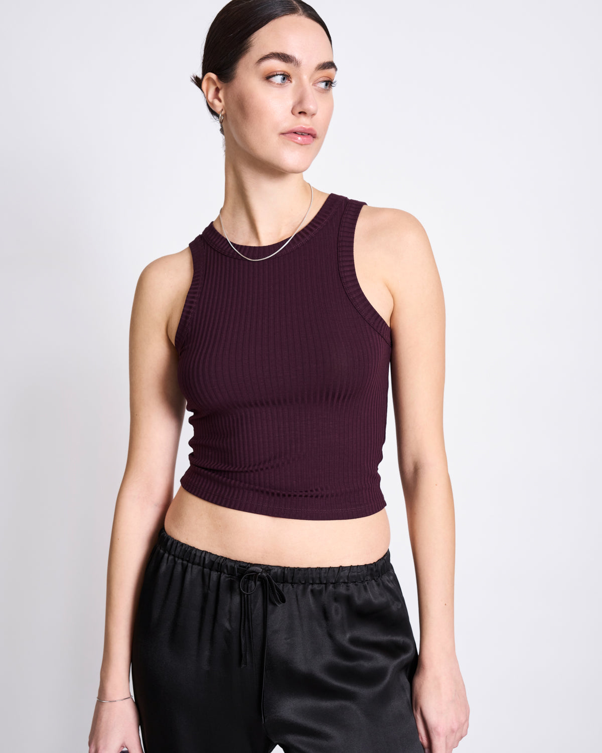 A woman with dark hair in a low bun wears the jan-n-june TENCEL™ LYOCELL RIB TANK TOP in Porto Cherry Lacquer and black satin pants, standing against a plain light background and looking slightly to the side.