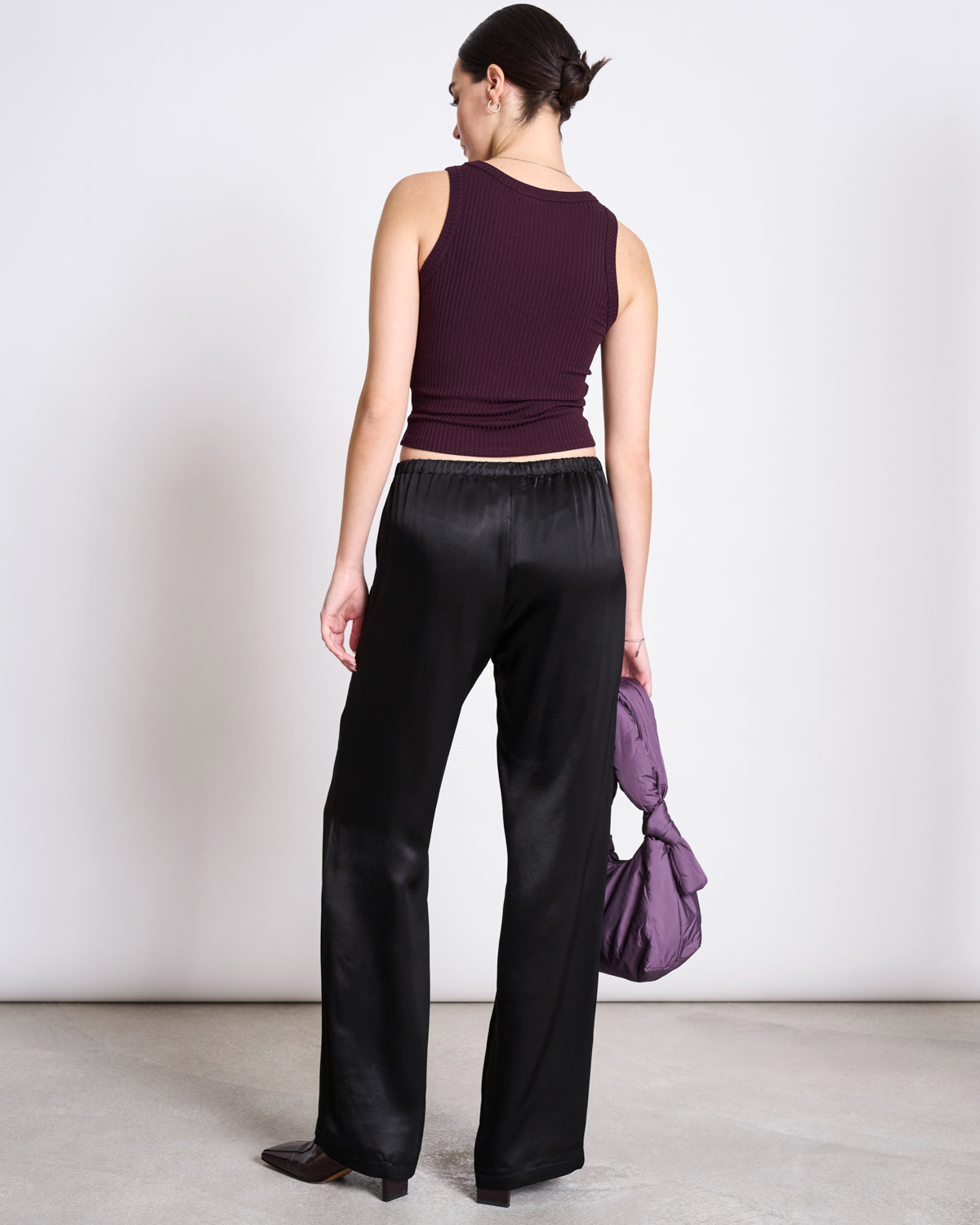 A woman with her hair tied back stands on a light floor against a plain wall, wearing jan-n-junes TENCEL™ LYOCELL RIB TANK TOP in Porto Cherry Lacquer and loose black satin pants, holding a matching purple handbag in her left hand.