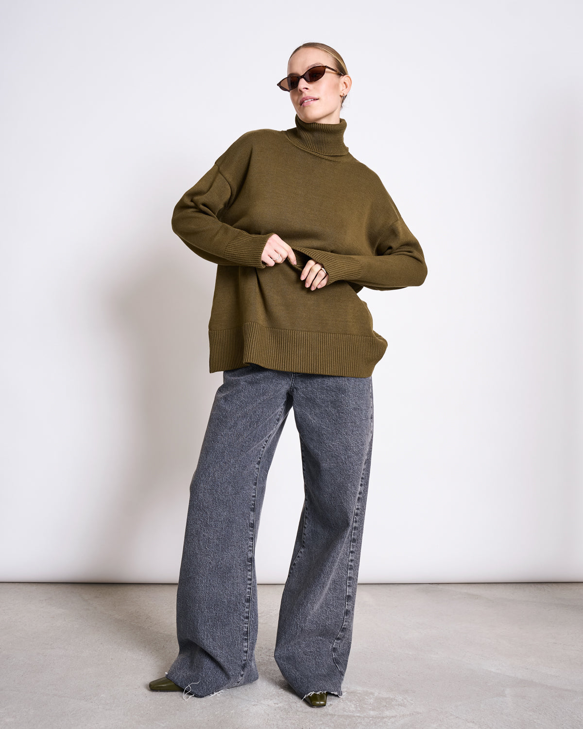 A person wearing jan-n-junes TURTLENECK MUA WOODY OLIVE GOTS, oversized in organic cotton, with wide-leg gray jeans and sunglasses, stands confidently against a plain white wall.