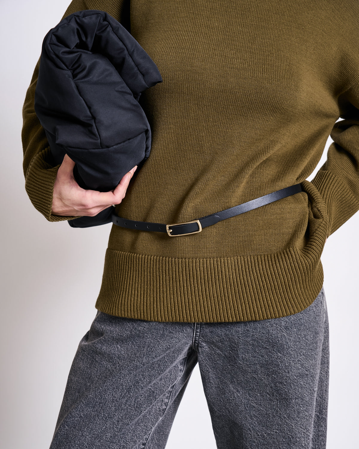 A person wearing the jan-n-june TURTLENECK MUA WOODY OLIVE GOTS jumper, grey jeans, and a slim black belt holds a black puffer jacket. Their face is not visible.