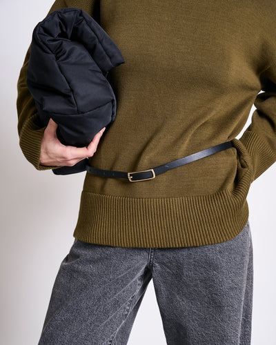 A person wearing the jan-n-june TURTLENECK MUA WOODY OLIVE GOTS jumper, grey jeans, and a slim black belt holds a black puffer jacket. Their face is not visible.