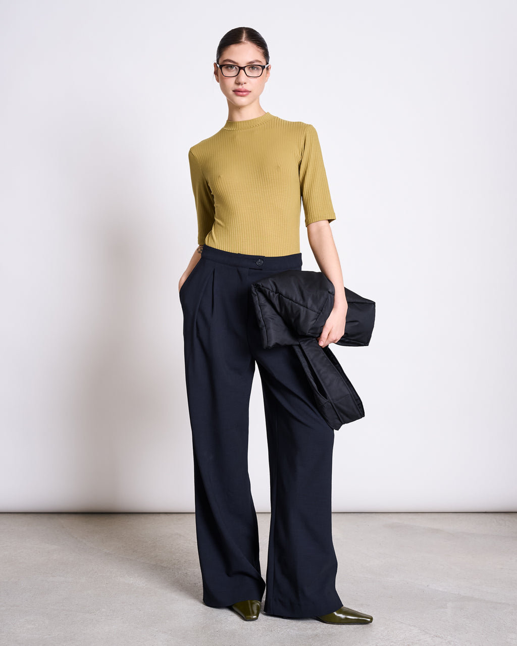A woman wearing the jan-n-june LYOCELL RIB T-SHIRT NINA in woody olive, navy wide-leg trousers, pointed green shoes, and glasses stands against a white wall, holding a black jacket with her hands in her pockets.