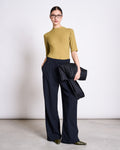 A woman wearing the jan-n-june LYOCELL RIB T-SHIRT NINA in woody olive, navy wide-leg trousers, pointed green shoes, and glasses stands against a white wall, holding a black jacket with her hands in her pockets.