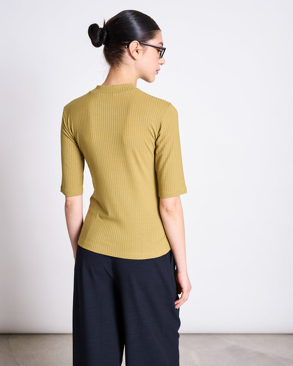 A woman with dark hair in a bun and glasses models the jan-n-june TENCEL™ LYOCELL RIB T-SHIRT NINA in woody olive, paired with loose black pants, standing with her back to the camera against a plain white background.