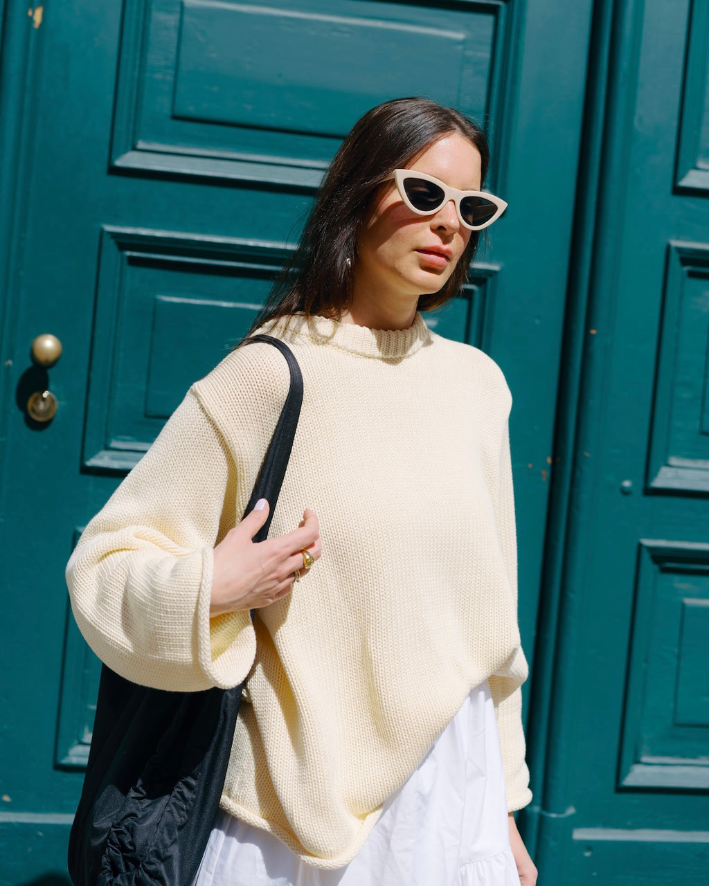 A woman stands in front of a teal door, wearing the jan-n-june JUMPER SONA BUTTERMILK GOTS (an oversized organic cotton sweater), a white skirt, cat-eye sunglasses, and holding a black tote bag on her shoulder.