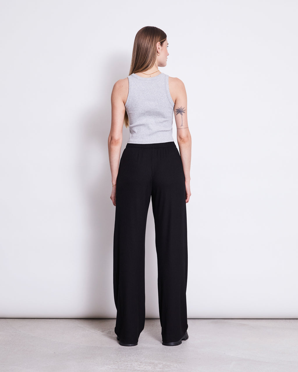 A woman with long straight hair and a tattoo on her upper left arm stands with her back to the camera, wearing jan-n-june’s ORGANIC COTTON TANK TOP PORTO LIGHT GREY GOTS and high-waisted black wide-leg pants.