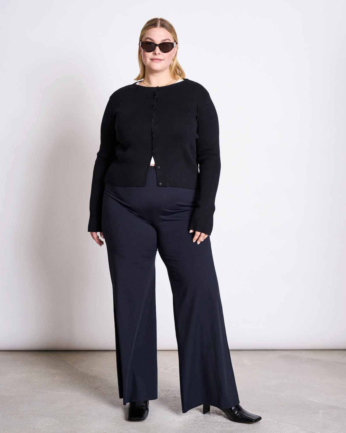A woman stands confidently against a plain white wall wearing black sunglasses, wide-leg pants, black boots, and the jan-n-june CREWNECK CARDIGAN GLIMT BLACK GOTS.