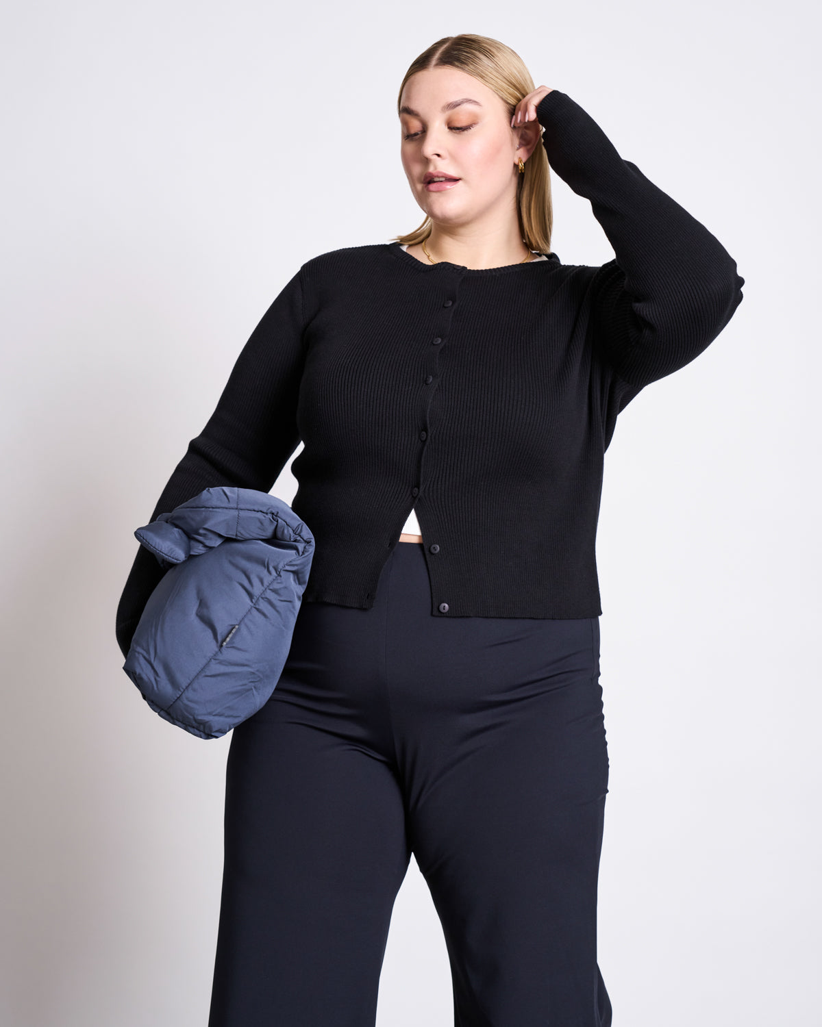 A woman in a jan-n-june CREWNECK CARDIGAN GLIMT BLACK GOTS and black pants holds a folded gray puffer jacket under her arm, touching her hair while standing against a plain light background.