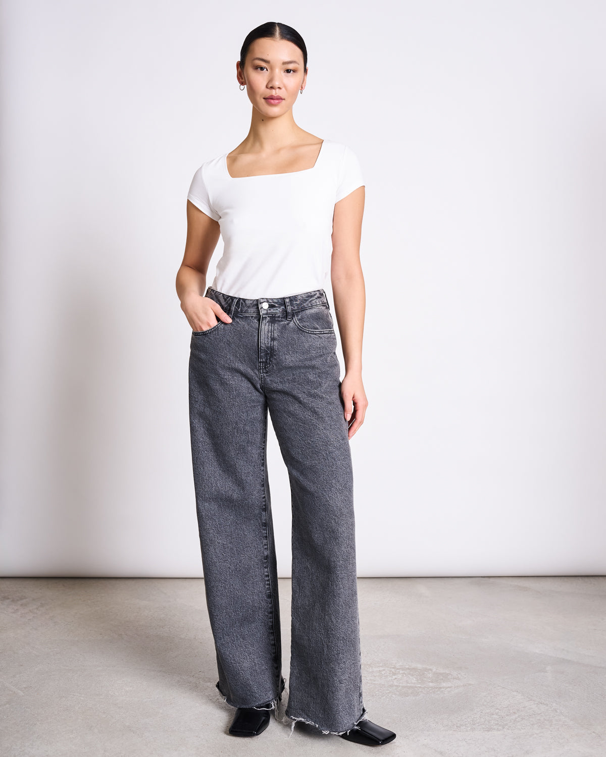 A person stands against a white background, wearing the jan-n-june T-SHIRT ARIMA WHITE GOTS with a square neck, paired with high-waisted wide-leg gray jeans and black shoes. One hand is in their pocket and they have a neutral expression.