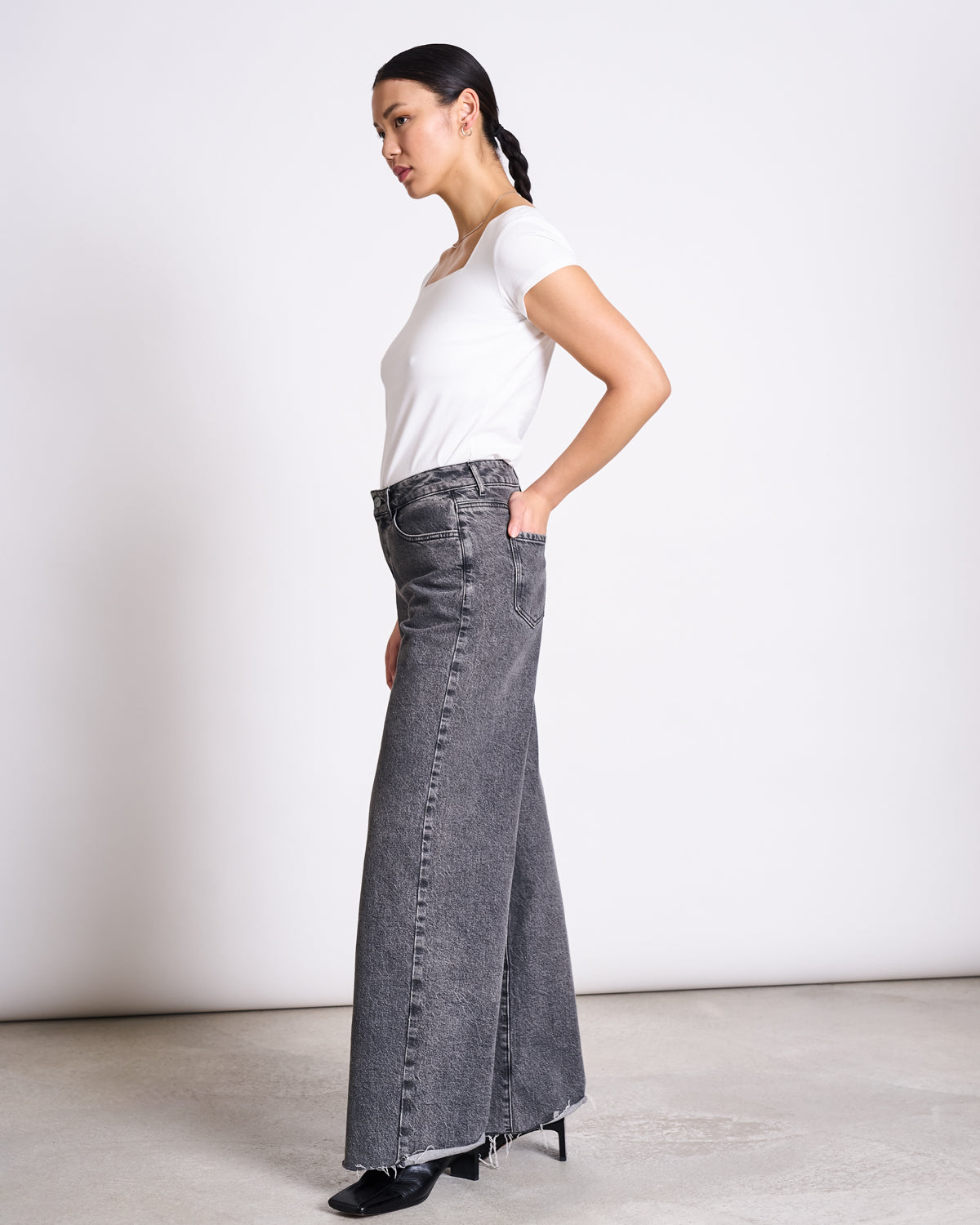 A woman stands sideways against a plain background, wearing jan-n-june’s T-SHIRT ARIMA WHITE GOTS with high-waisted wide-leg gray jeans and black pointed shoes. Her hair is styled back in a braid.