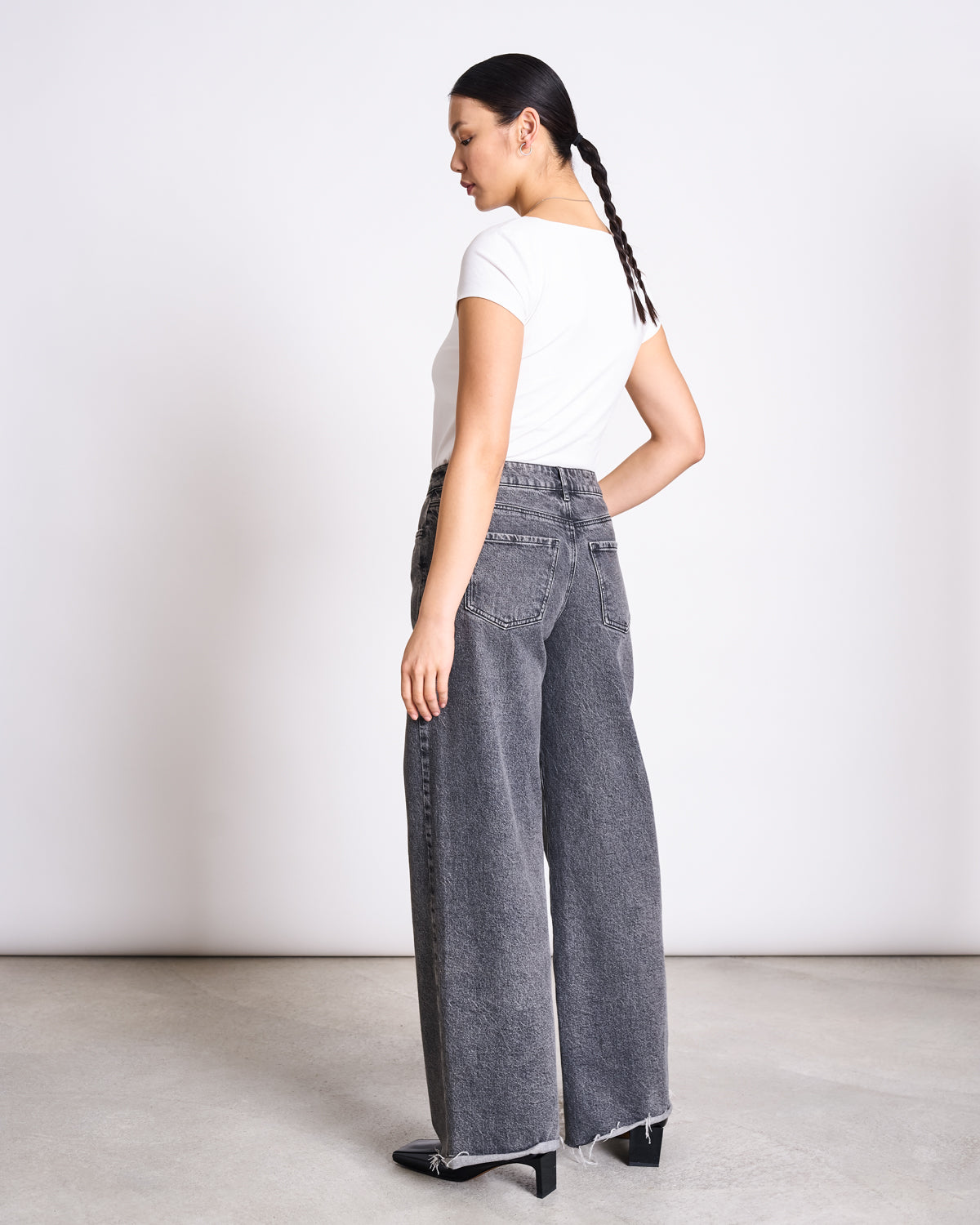 A woman with a long braid wears the jan-n-june T-SHIRT ARIMA WHITE GOTS and high-waisted, wide-leg gray jeans with frayed hems, standing sideways and looking over her shoulder against a plain background.